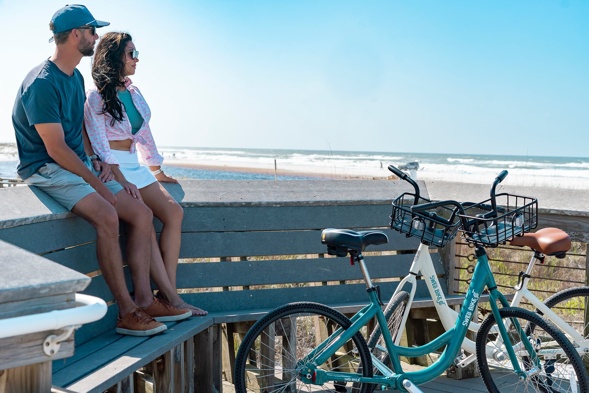 A couple on a boardwalk bench above the beach, Swell bikes parked beside them